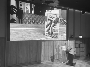 Kelly speaking on stage in front of a large screen above her head. It is at Bletchley Park.