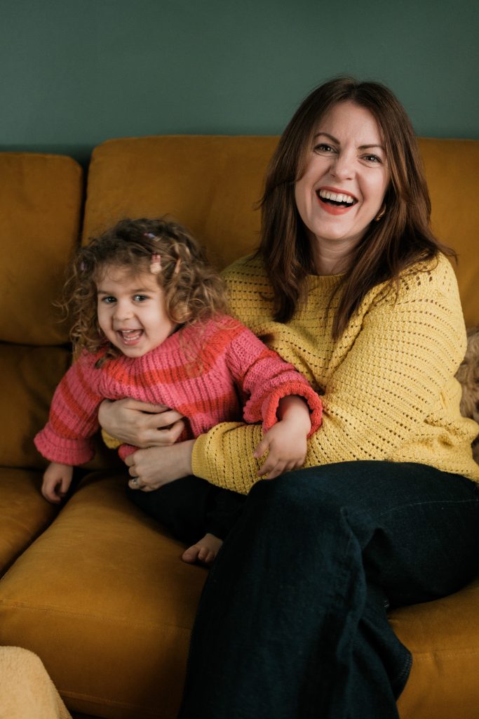 Kelly sits on on a mustard sofa wearing jeans and a yellow jumper, her daughter sits on her lap wearing a pink jumper.